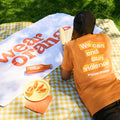 A boy lounges on a picnic blanket wearing an orange short-sleeve unisex tee that reads: We can end gun violence #wearorange. A white towel with the Wear Orange logo in orange print is draped over the picnic blanket. An orange stainless steel water bottle with a white Wear Orange logo lays atop the towel, along with a plate of orange slices.