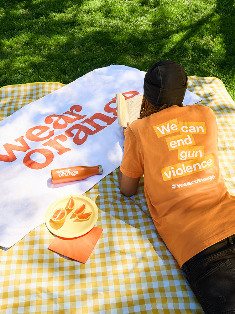 A boy lounges on a picnic blanket wearing an orange short-sleeve unisex tee that reads: We can end gun violence #wearorange. A white towel with the Wear Orange logo in orange print is draped over the picnic blanket. An orange stainless steel water bottle with a white Wear Orange logo lays atop the towel, along with a plate of orange slices.