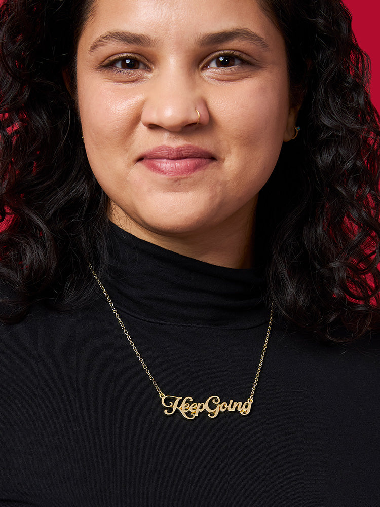 Woman wearing a gold name plate style necklace with the phrase Keep Going in script font.