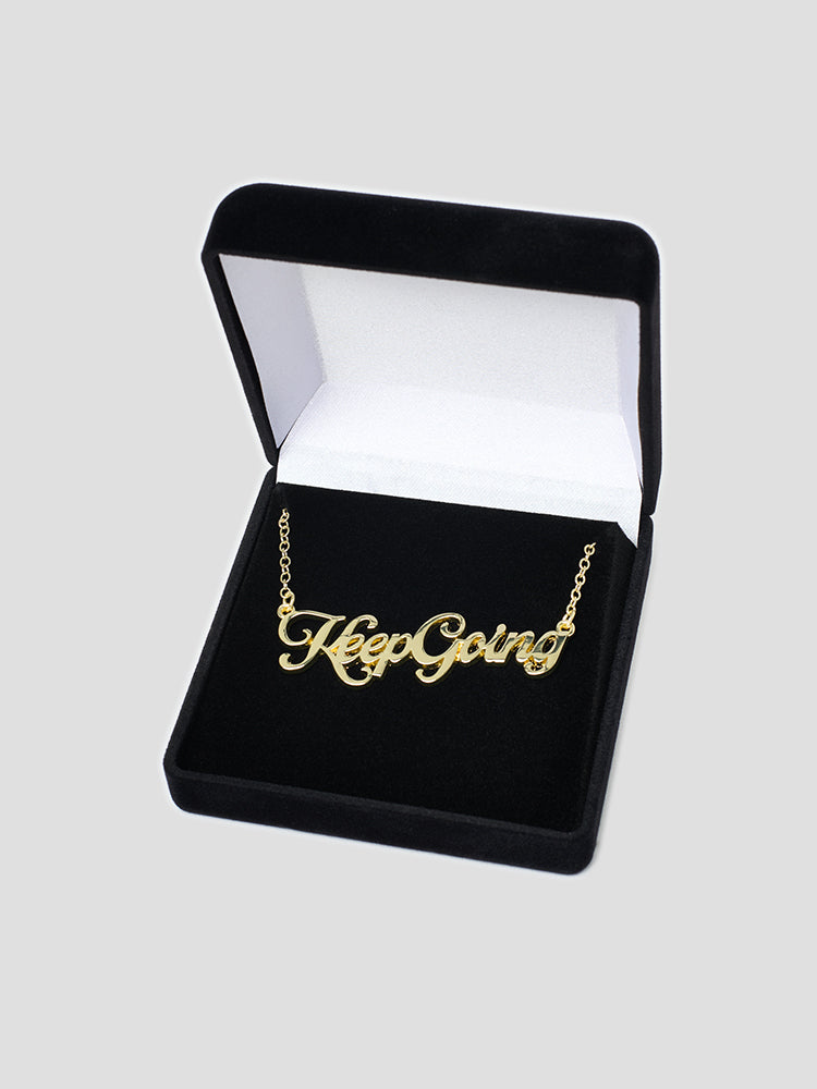 A gold name plate style necklace with the phrase Keep Going in script font in a black velvet box.