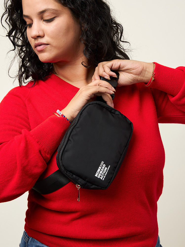Woman wearing an all black nylon belt bag with an adjustable strap, zip closure, and small white Moms Demand Action Logo printed on the bottom left corner.