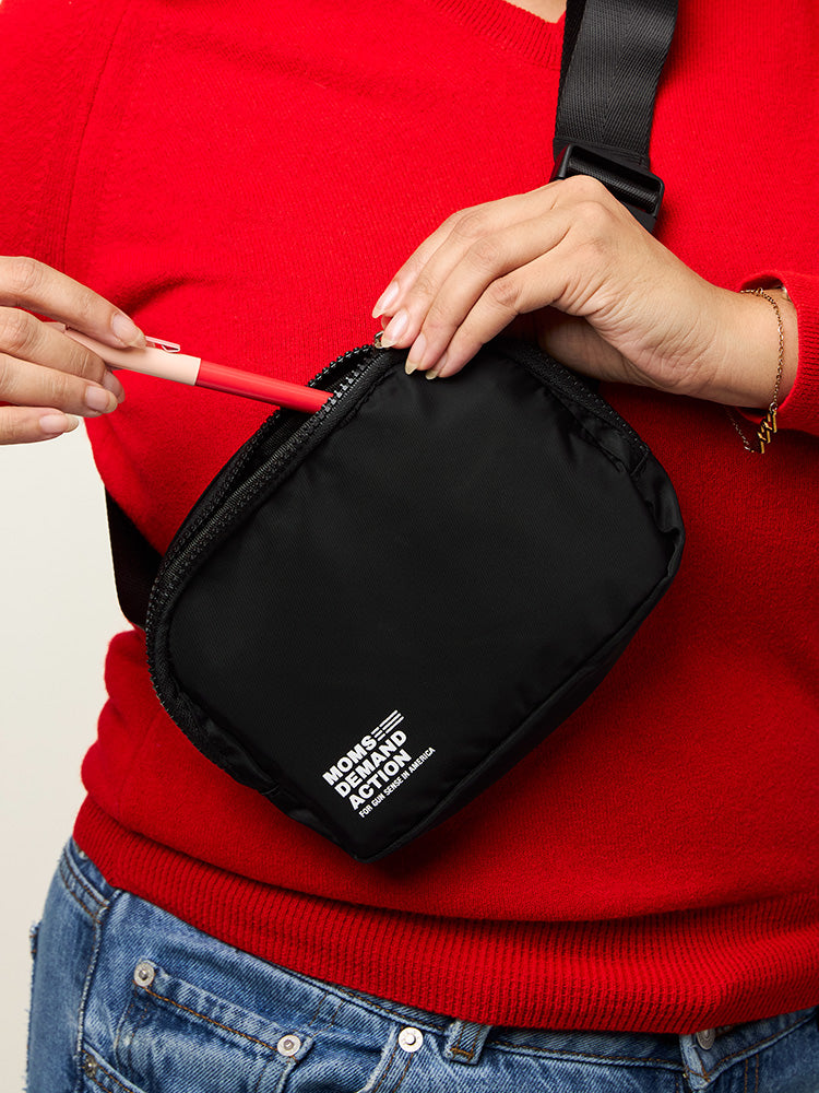 Woman pulling pen out of an all black nylon belt bag with an adjustable strap, zip closure, and small white Moms Demand Action Logo printed on the bottom left corner.