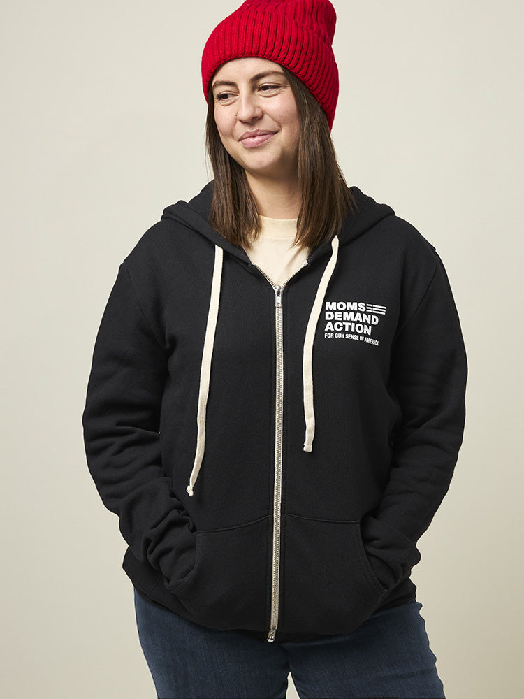 Front view of woman wearing a black zip-up hooded sweatshirt with two front pockets and a small white Moms Demand Action logo printed on left chest.