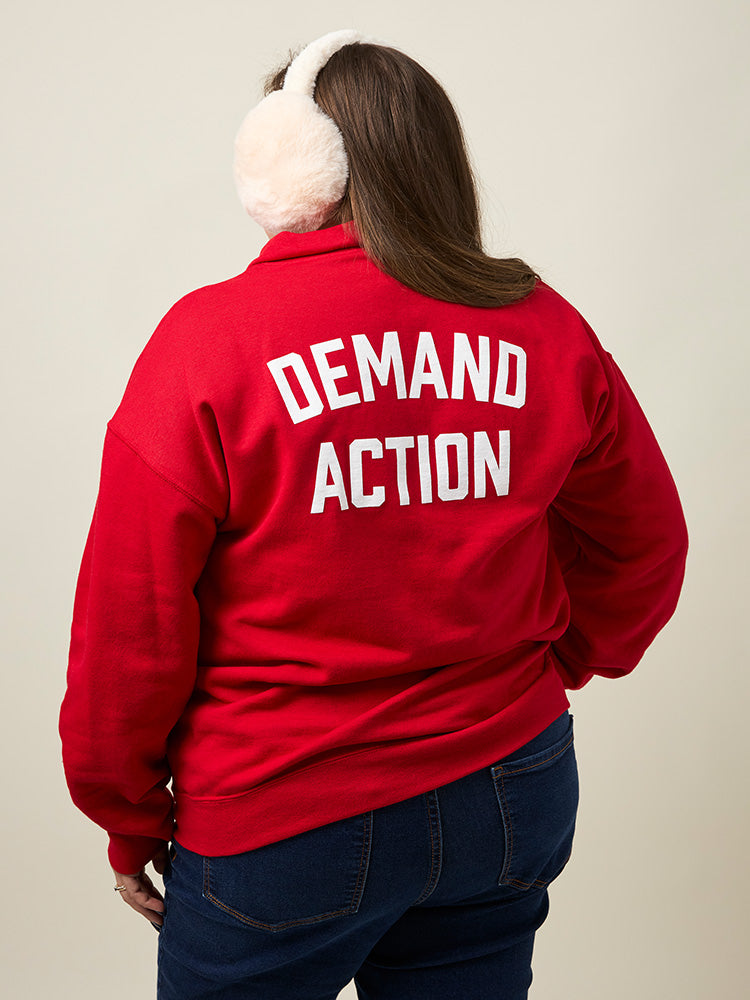 Back view of woman wearing a red quarter-zip collared sweatshirt with the phrase DEMAND ACTION printed in large white all caps text across the back.