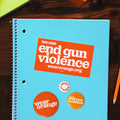 Notebook on desk surrounded by office supplies. Rectangular orange sticker is stuck to the notebook with white text that reads: we can end gun violence wearorange.org. Three circular buttons of different sizes. Largest button is dark orange with a white Wear Orange logo. Medium button is light orange with white text that reads: we can end gun violence. Smallest button is white with orange text that reads: wear orange.