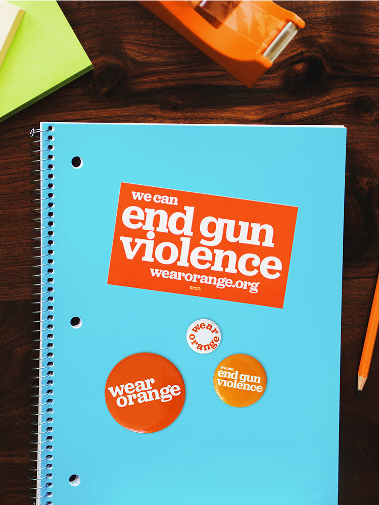 Notebook on desk surrounded by office supplies. Rectangular orange sticker is stuck to the notebook with white text that reads: we can end gun violence wearorange.org. Three circular buttons of different sizes. Largest button is dark orange with a white Wear Orange logo. Medium button is light orange with white text that reads: we can end gun violence. Smallest button is white with orange text that reads: wear orange.