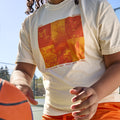 Cream colored short-sleeve unisex t-shirt, front view. There is a 2x3 grid with orange duotone images of candles, birds flying, lanterns in the sky, marigolds, a fist raised, and a monarch butterfly. In small, orange all-caps print, it reads: Wear orange for a future free from gun violence.