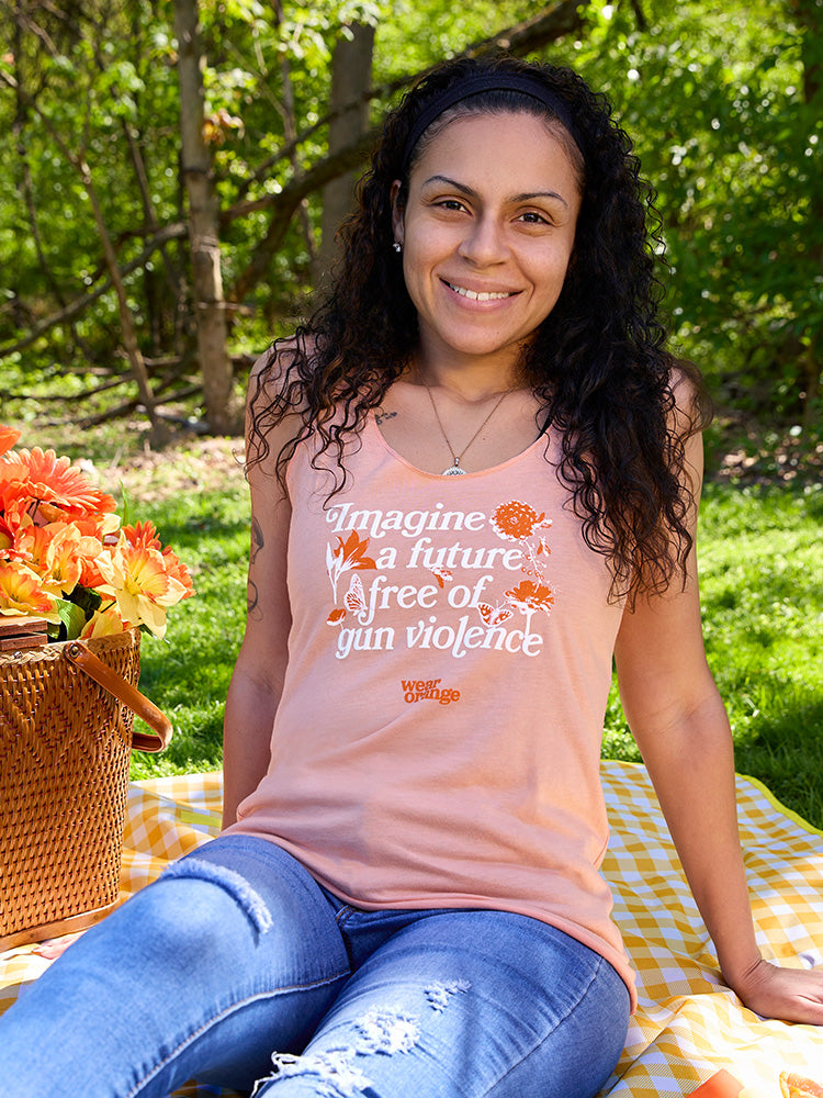 A woman smiles wearing a peach colored women's cut racerback tank top. In an italic, serif font, it reads: Imagine a future free of gun violence. The text is embellished with orange and white flowers, butterflies, and bees. A small Wear Orange logo is printed in orange below the text.