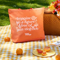 Orange tote bag with orange straps and a zipper enclosure leans against a wicker picnic basket atop a yellow gingham picnic blanket, scattered with oranges and a book. In an italic, serif font, it reads: Imagine a future free of gun violence. The white text is embellished with light orange and white flowers, butterflies, and bees. A small Wear Orange logo is printed in white below the text.
