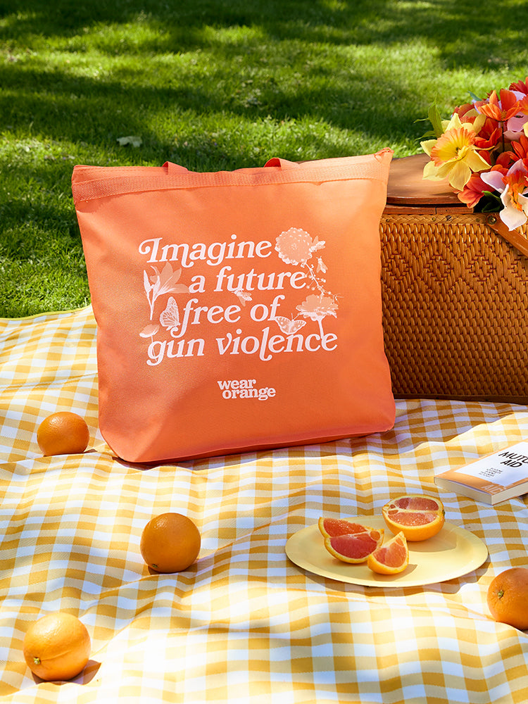 Orange tote bag with orange straps and a zipper enclosure leans against a wicker picnic basket atop a yellow gingham picnic blanket, scattered with oranges and a book. In an italic, serif font, it reads: Imagine a future free of gun violence. The white text is embellished with light orange and white flowers, butterflies, and bees. A small Wear Orange logo is printed in white below the text.