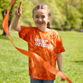 A girl wearing an orange tie-dye short-sleeve unisex kids t-shirt with a white Wear Orange logo printed across the chest smiles as she twirls an orange ribbon in a grassy field.