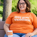 A woman sits on a park bench wearing an orange t-shirt that reads: we can end gun violence. She is holding a white ceramic mug with the Wear Orange logo printed in orange.
