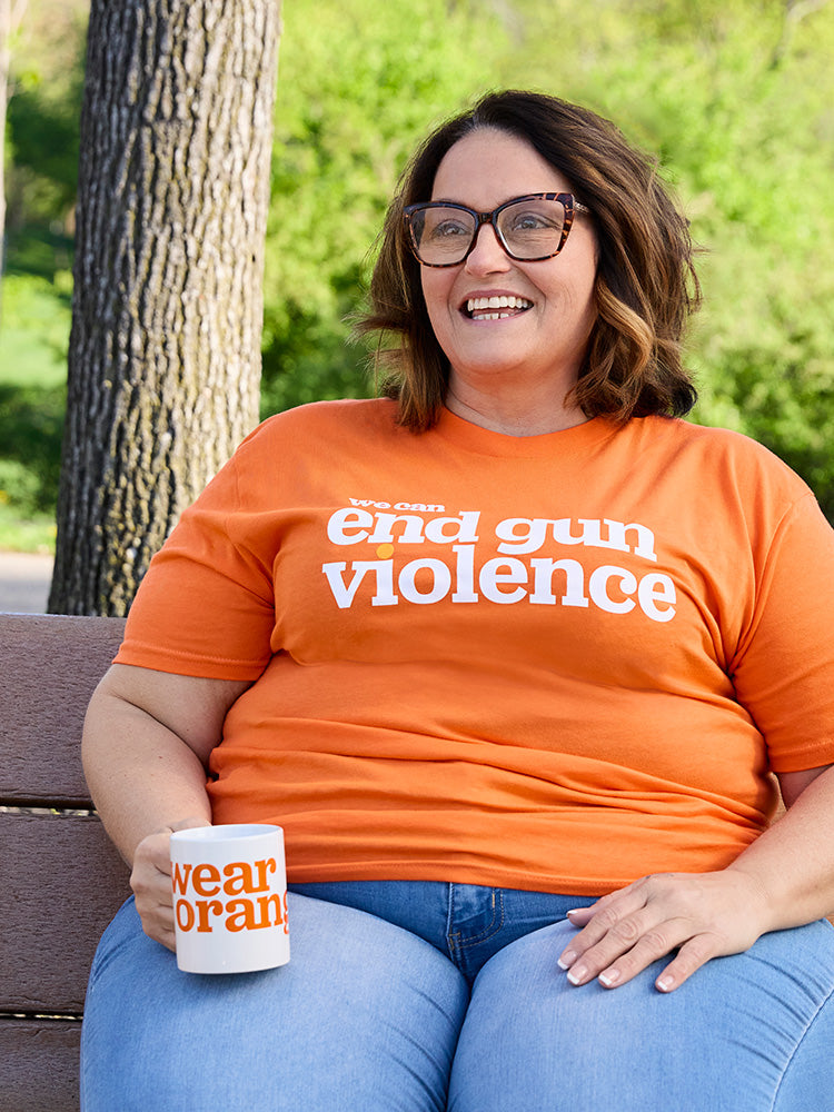 A woman sits on a park bench wearing an orange t-shirt that reads: we can end gun violence. She is holding a white ceramic mug with the Wear Orange logo printed in orange.