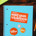Notebook on desk surrounded by office supplies. Rectangular orange sticker is stuck to the notebook with white text that reads: we can end gun violence wearorange.org. Three circular buttons of different sizes. Largest button is dark orange with a white Wear Orange logo. Medium button is light orange with white text that reads: we can end gun violence. Smallest button is white with orange text that reads: wear orange.
