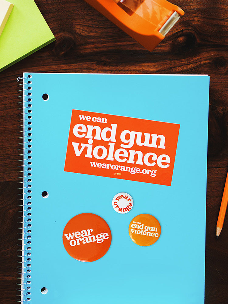 Notebook on desk surrounded by office supplies. Rectangular orange sticker is stuck to the notebook with white text that reads: we can end gun violence wearorange.org. Three circular buttons of different sizes. Largest button is dark orange with a white Wear Orange logo. Medium button is light orange with white text that reads: we can end gun violence. Smallest button is white with orange text that reads: wear orange.