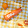 An orange stainless steel water bottle with a silver screw-on lid lays atop a yellow gingham picnic blanket, surrounded by oranges. A white Wear Orange logo is printed vertically on the bottle.
