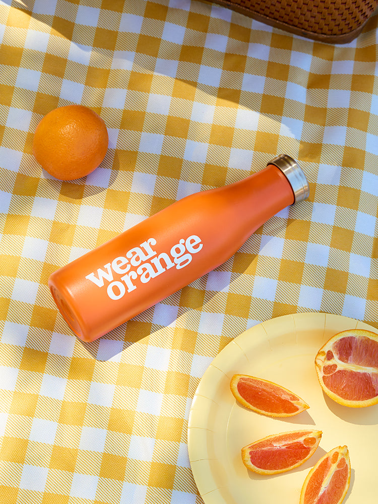An orange stainless steel water bottle with a silver screw-on lid lays atop a yellow gingham picnic blanket, surrounded by oranges. A white Wear Orange logo is printed vertically on the bottle.