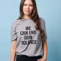 Front view of woman wearing a light gray unisex short sleeve t-shirt with the text WE CAN END GUN VIOLENCE printed in black all caps.